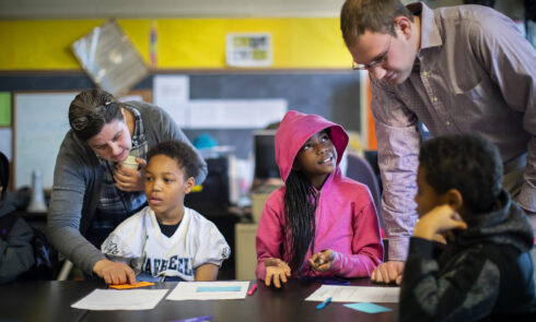 Two teachers speak to children at a desk in a classroom.