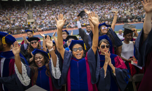 A group of graduate students in caps and gowns cheer at their commencement ceremony.
