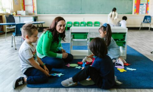 Three children and a schoolteacher sit in a circle in a classroom.