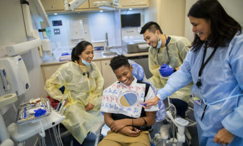 Three dentists smile as a young client joyfully looks at his teeth post-routine checkup.