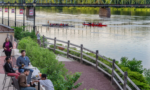 Groups of people sit at tables by a river.