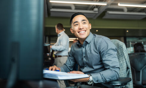 A young man smiles while working at his desk in an office.