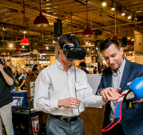 A man wearing a virtual reality headset is aided by a man in a suit.
