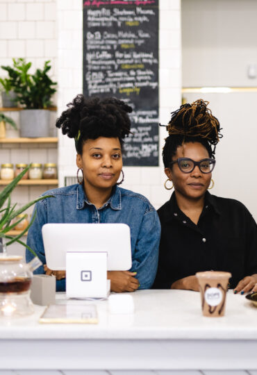 Two women pose for the camera behind the counter of a small retail store.